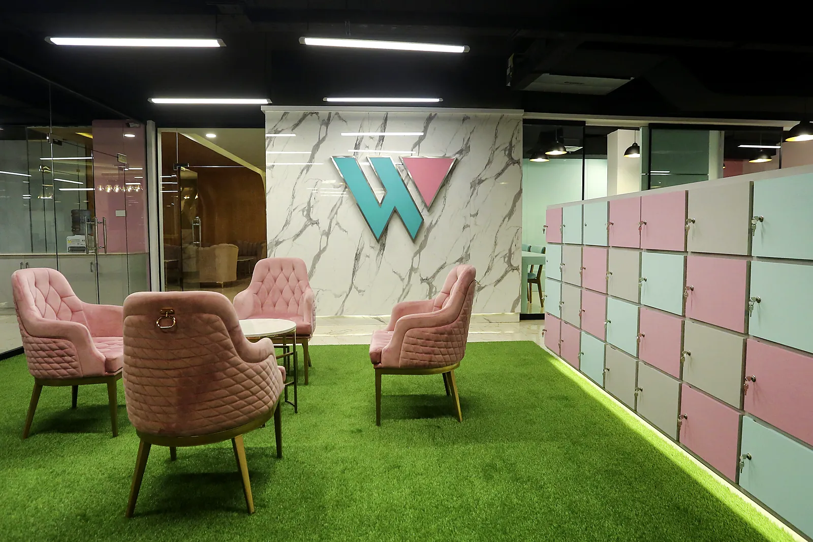 Community lounge with pink armchairs, green turf and member lockers at TheWing.pk